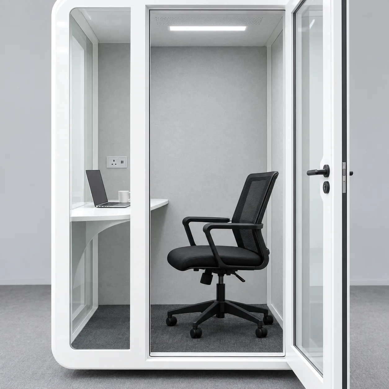 Inside a white Slope Pod: pale acoustic walls, bright LED light, a built in desk with a laptop and coffee, and a mesh chair